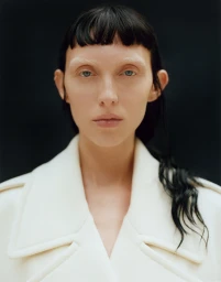 Close-up portrait of a woman with bleached eyebrows and dark hair wearing an off-white coat against a dark background