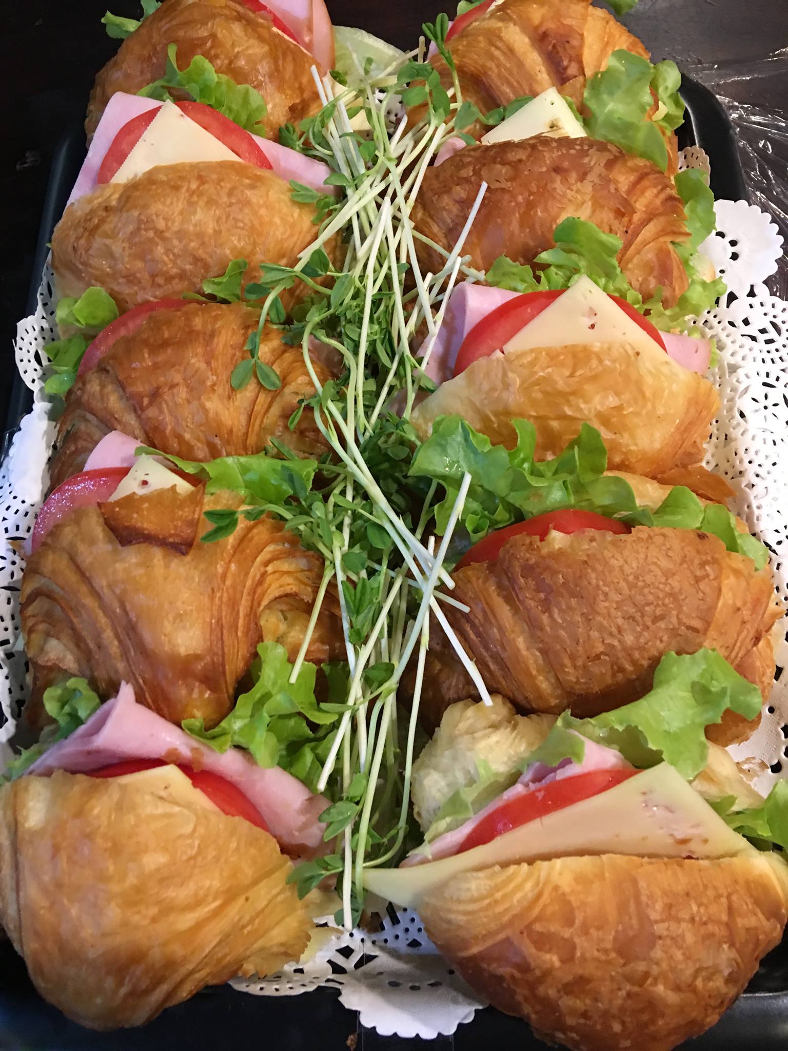 A platter of fresh croissant sandwiches filled with ham, cheese, lettuce, tomato, and sprouts, perfect for a light and tasty meal or snack.