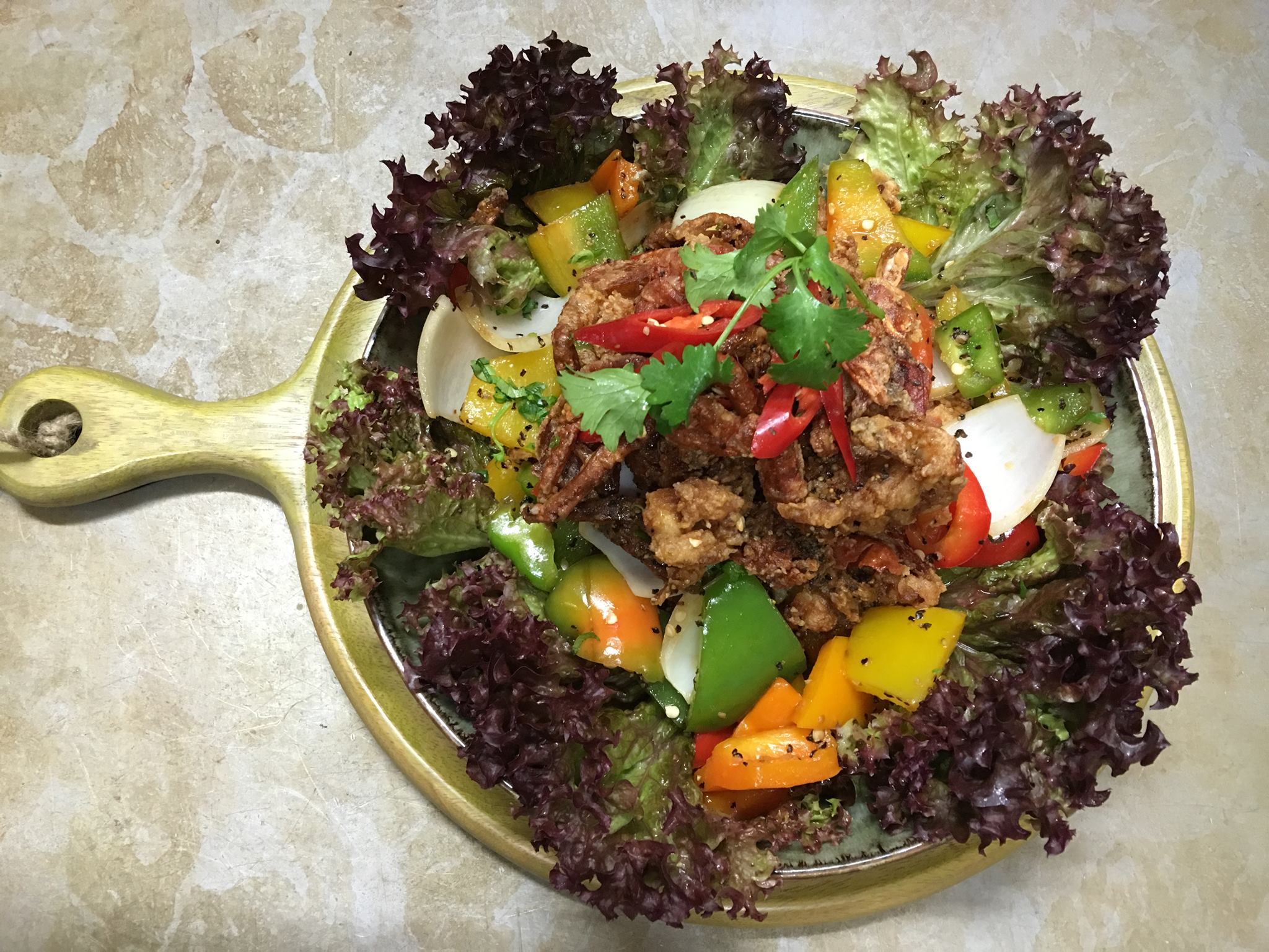 Overhead shot of a wooden serving board with a salad, featuring fried crab, colorful bell peppers, onions, cilantro, and red lettuce.