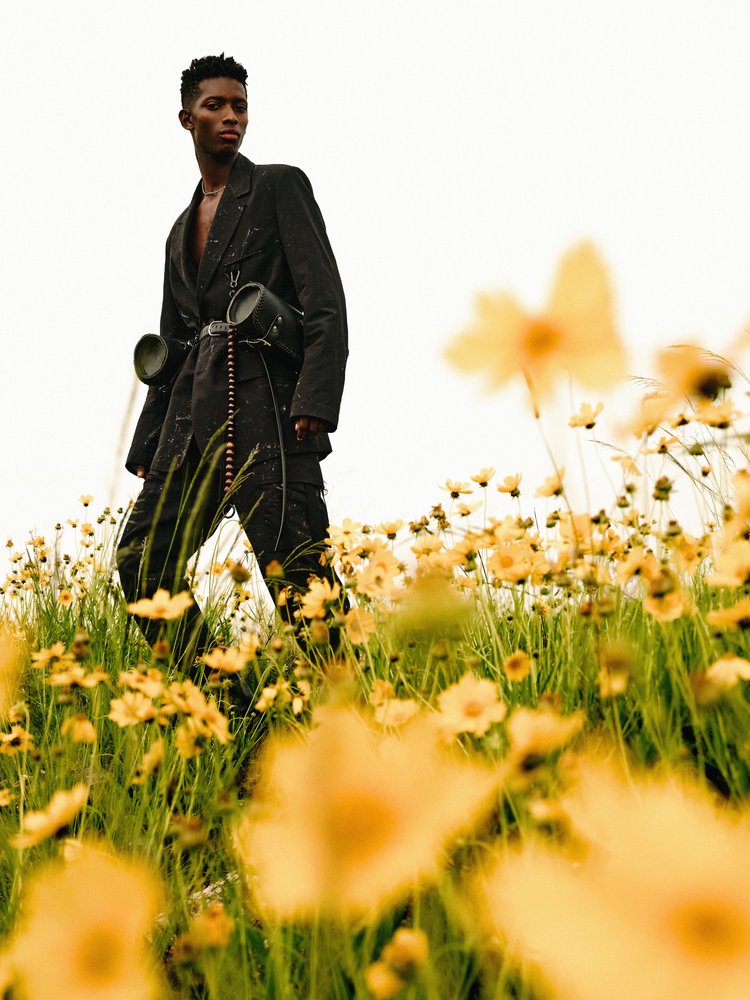 Man in an untied suit with speaker-like accessories on a belt stands in a field of Coreopsis flowers against a white sky outdoors.