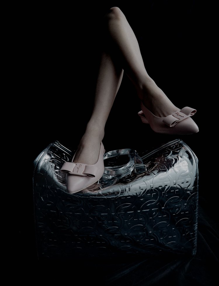 Pale pink high-heeled shoes with bow detail rest on a reflective silver handbag against a stark black backdrop.