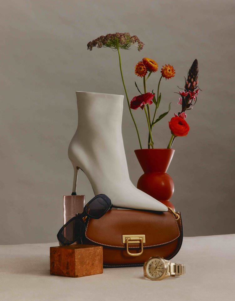 Fashion still life featuring a white high-heeled boot, purse, sunglasses, lipstick, flowers in a vase, and jewelry accessories.