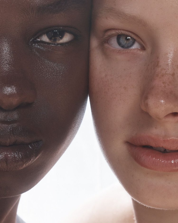Close-up portrait of two women, one with very dark skin and one with very light skin, their faces juxtaposed against a white background.