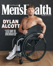 Dylan Alcott seated in a wheelchair wearing dark athletic shorts and sneakers against a textured dark wall.