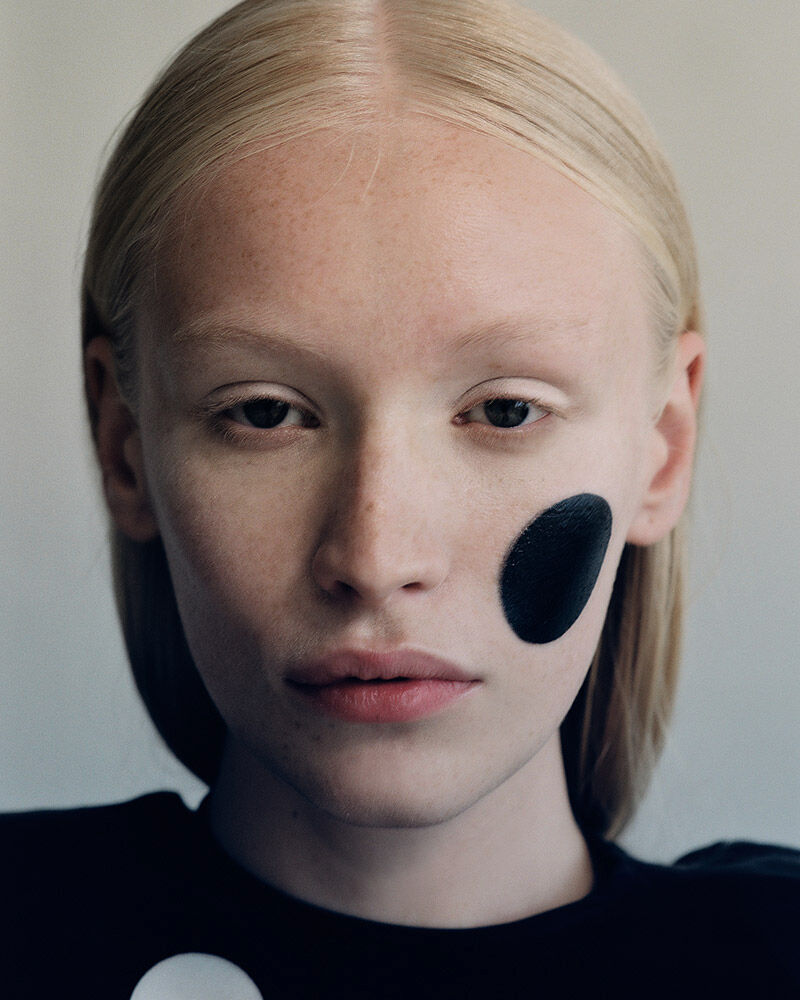 Close-up portrait of a fair-skinned adult female with blonde hair and a black dot on her cheek, wearing a black and white polka dot top.