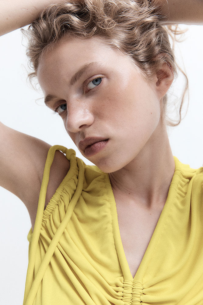 Close-up portrait of a blonde woman with blue eyes wearing a yellow v-neck top, posing with her hands behind her head against a white background.