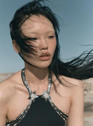 Close-up portrait of an East Asian woman with dark hair blowing in the wind wearing a black halter top with silver star embellishments against a clear sky