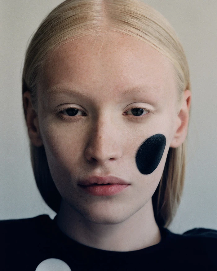 Close-up beauty portrait of a blonde model with a circular black cosmetic patch on her cheek.