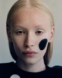 Close-up beauty portrait of a blonde model with a circular black cosmetic patch on her cheek.