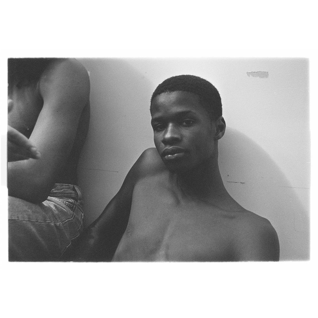 Black and white portrait of a young adult man with dark hair, with another person partially visible in the background.