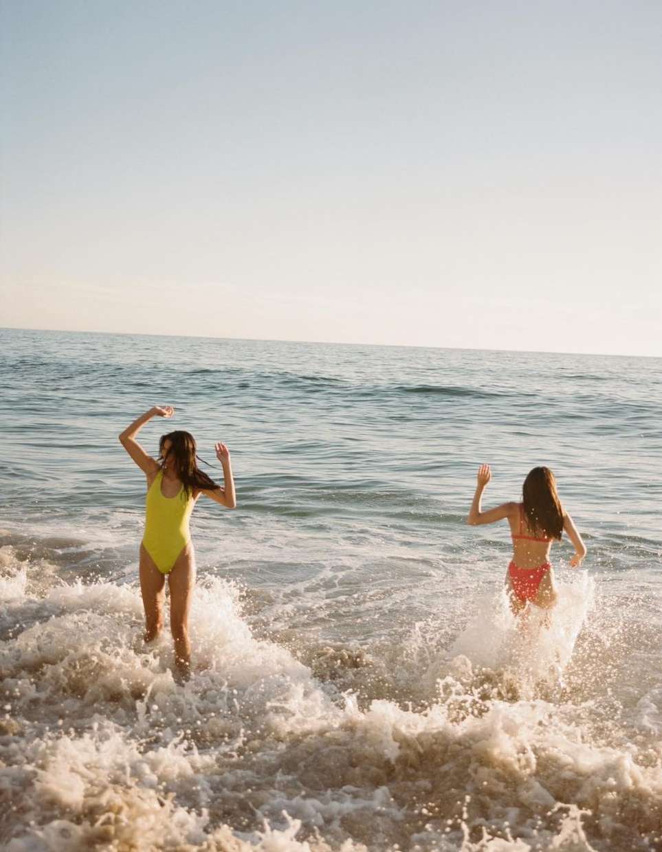 Two women in swimsuits joyfully wade into the ocean on a sunny day, captured at eye-level with splashing water and blue sky.