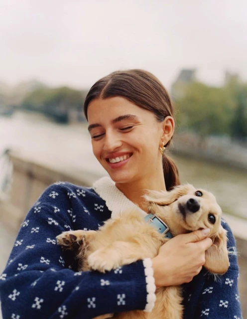 Smiling woman with brown hair in blue patterned sweater holds a tan dachshund puppy outdoors along a stone wall with a river backdrop.