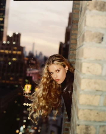 A blonde woman leaning out from a brick building exterior looking toward the camera at dusk