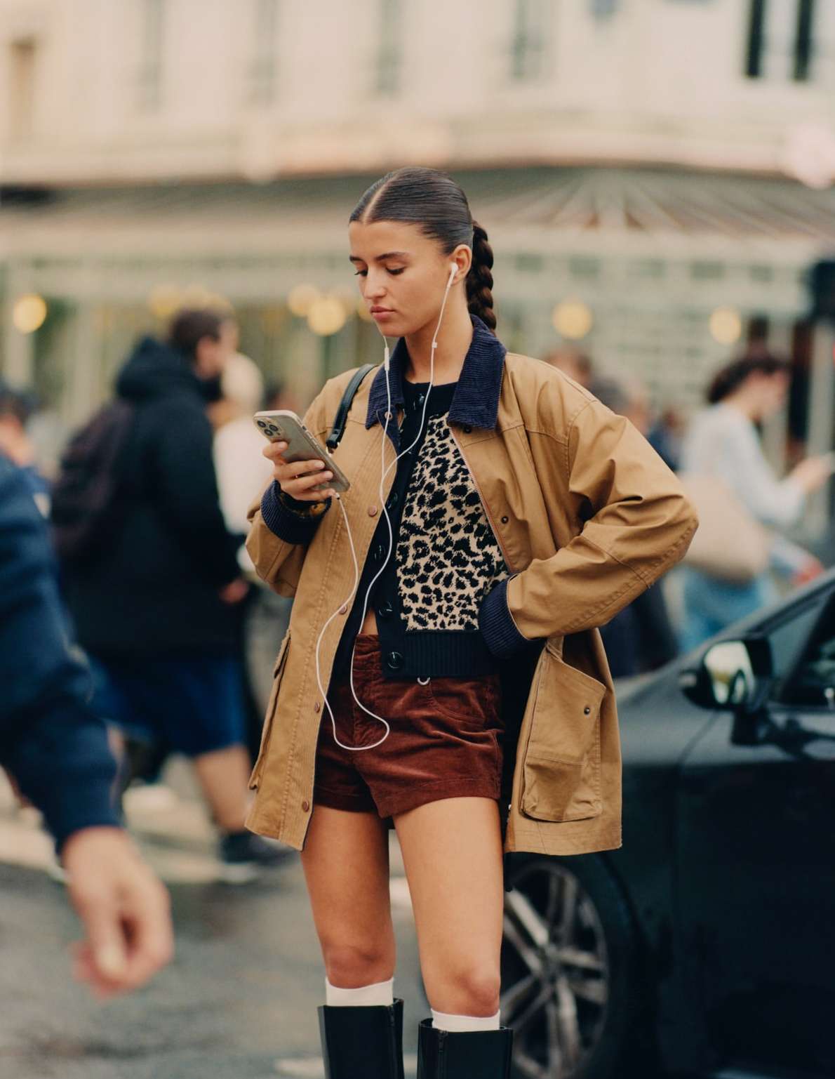 A young woman in a tan jacket uses her smartphone while wearing wired headphones on a city street with pedestrians and cars.
