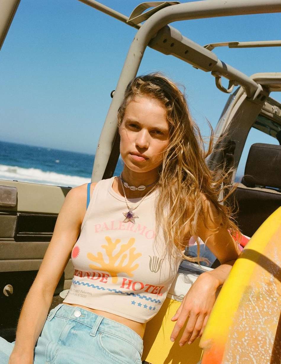 Woman in cream tank top and light blue jeans seated inside an open jeep with a surfboard and ocean view by Ryan Brabazon.