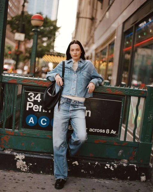A young adult woman in a denim jacket and jeans poses next to a subway entrance in an urban setting with buildings in the background.