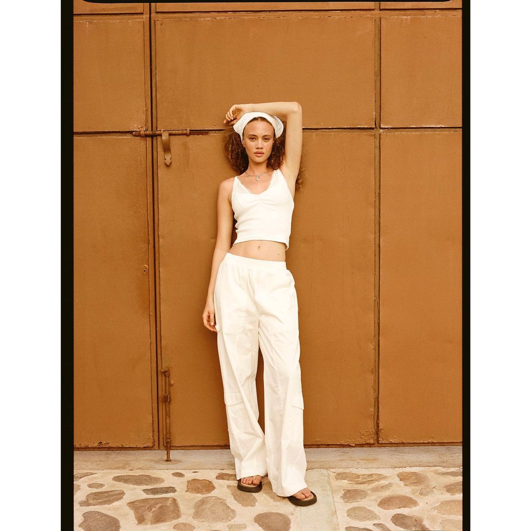 Woman in white top and pants poses against a brown wall with stone ground. The shot is well-lit with a shallow depth of field.