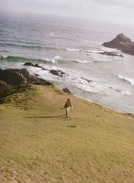 Woman in casual clothes walking on green hilltop near ocean with crashing waves, under an overcast sky, creating a natural, serene view.