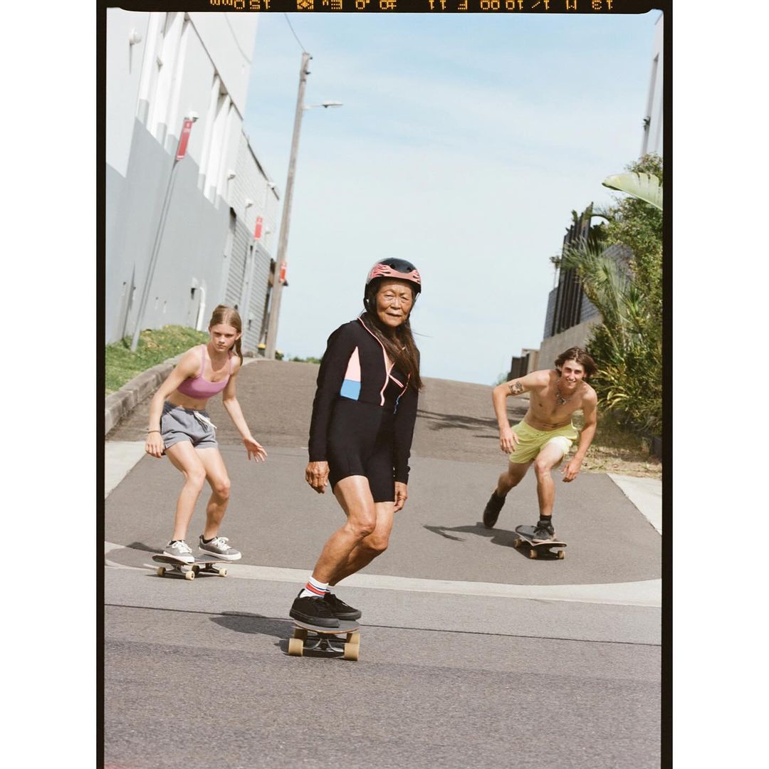 Three people skateboarding on an asphalt road. The person in the foreground wears a helmet and dark clothing, smiling confidently.