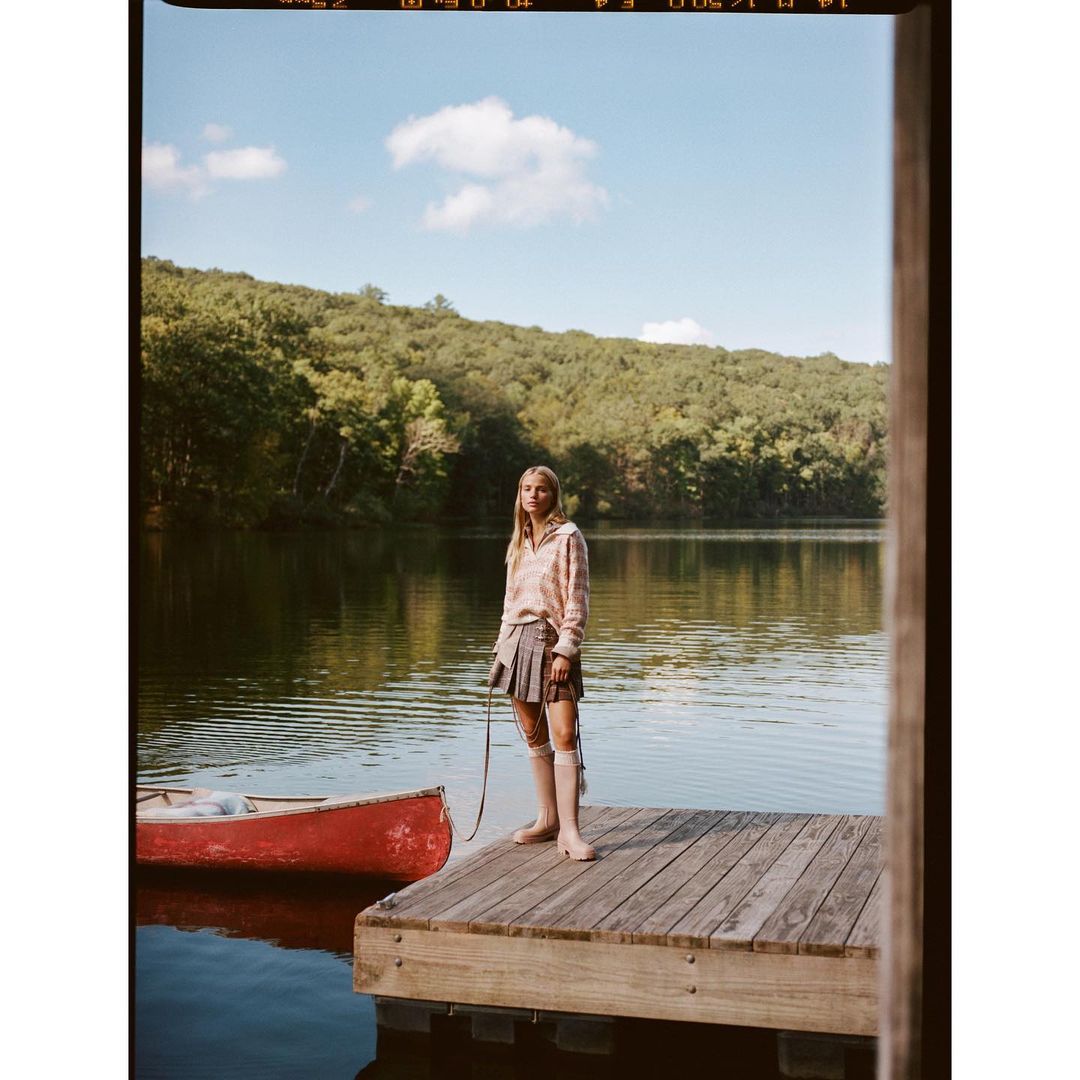 Young woman in preppy fashion stands on a wooden dock by a lake with a red canoe, surrounded by trees and a bright sky.