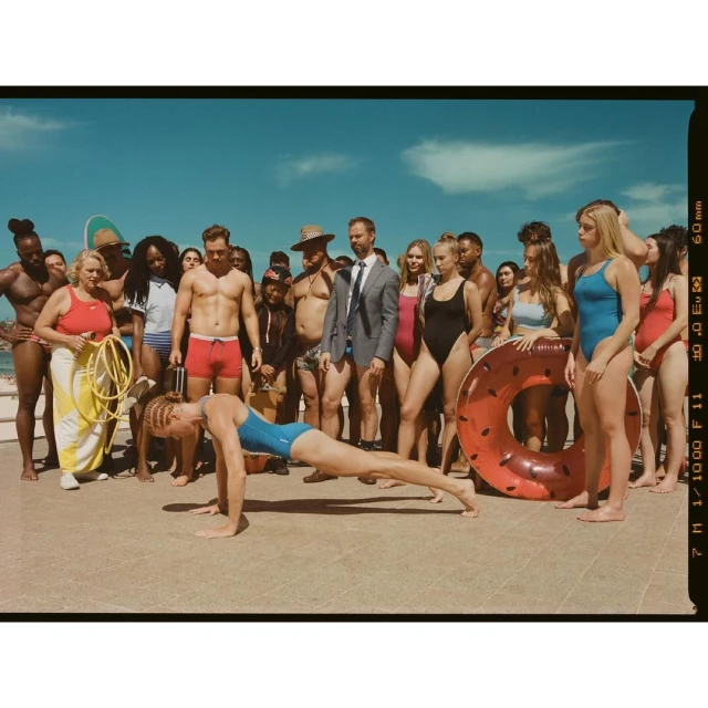 Outdoor shot of diverse group on concrete with a woman doing push-ups in a blue bathing suit in front, ocean and sky background.