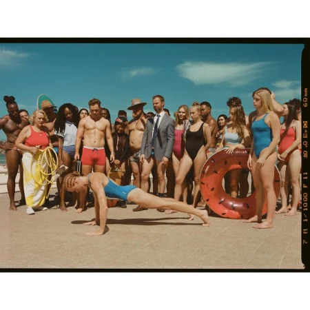 Outdoor shot of diverse group on concrete with a woman doing push-ups in a blue bathing suit in front, ocean and sky background.