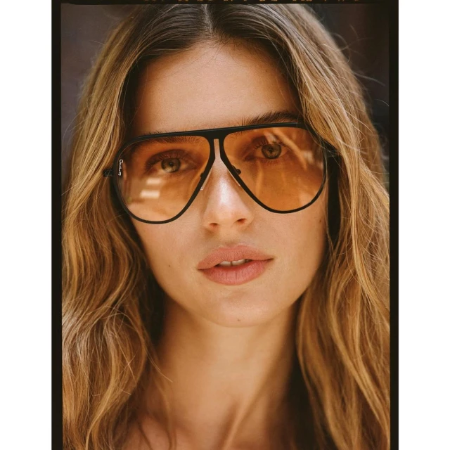 Close-up portrait of a young adult woman with long wavy hair wearing aviator sunglasses with orange lenses and a black frame.