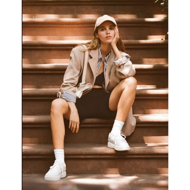 Young woman in an oversized blazer and baseball cap sitting on outdoor steps with a white purse, white socks, and white sneakers.