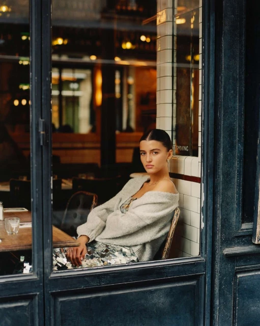 A young woman is sitting at a table inside a cafe, wearing a grey sweater and patterned dress, as seen through a window during daytime.