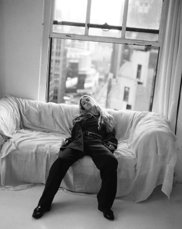 Black and white portrait of a woman lounging on a sofa by a window overlooking a cityscape, wearing a jacket and pants.