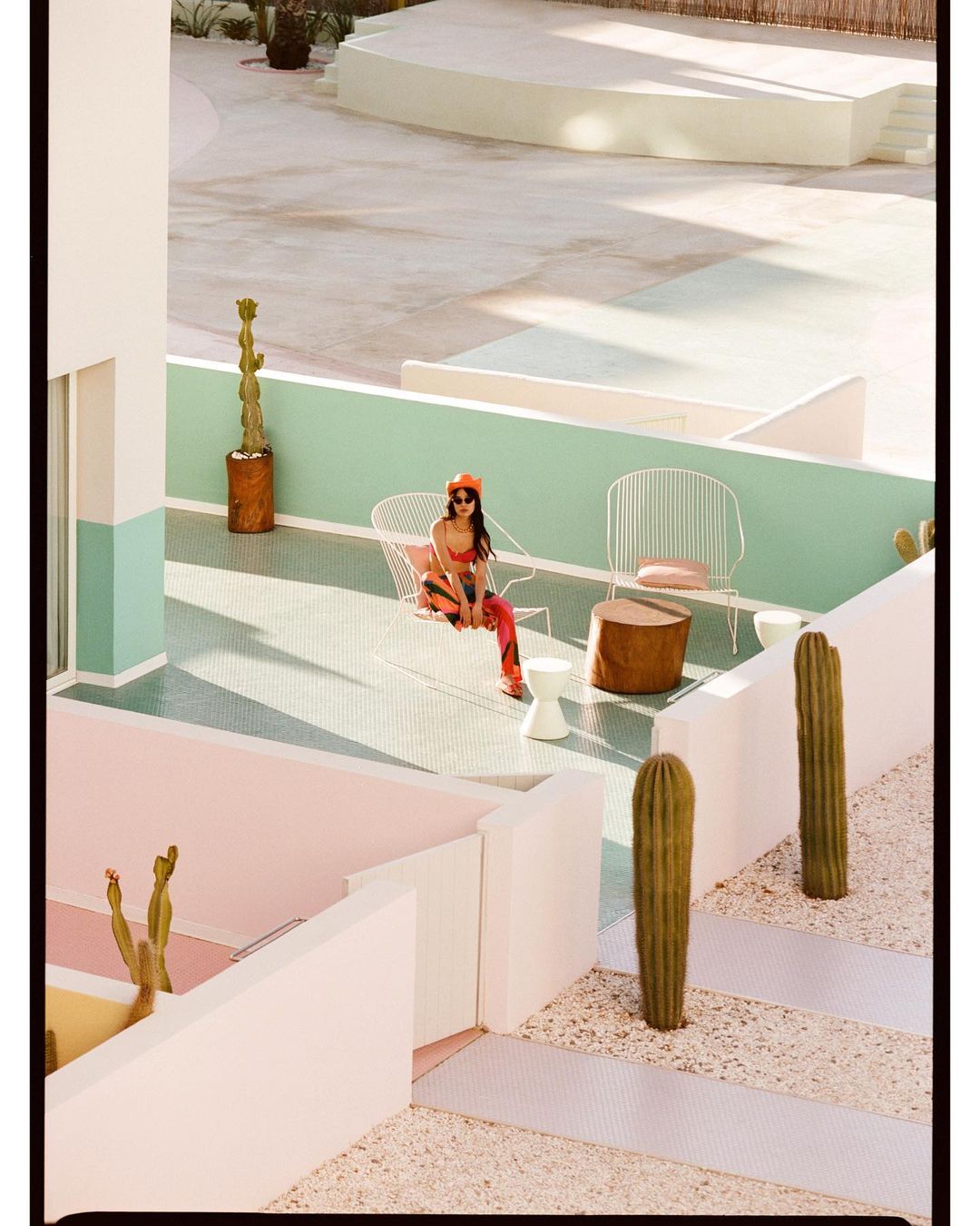 A woman in a vibrant orange outfit lounges in a white wire chair on a colorful patio, complemented by cacti, creating a modern aesthetic.