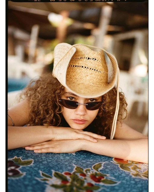 Indoor portrait of a young adult woman with curly hair, wearing sunglasses and a cowboy hat, posing with hands on a floral table.