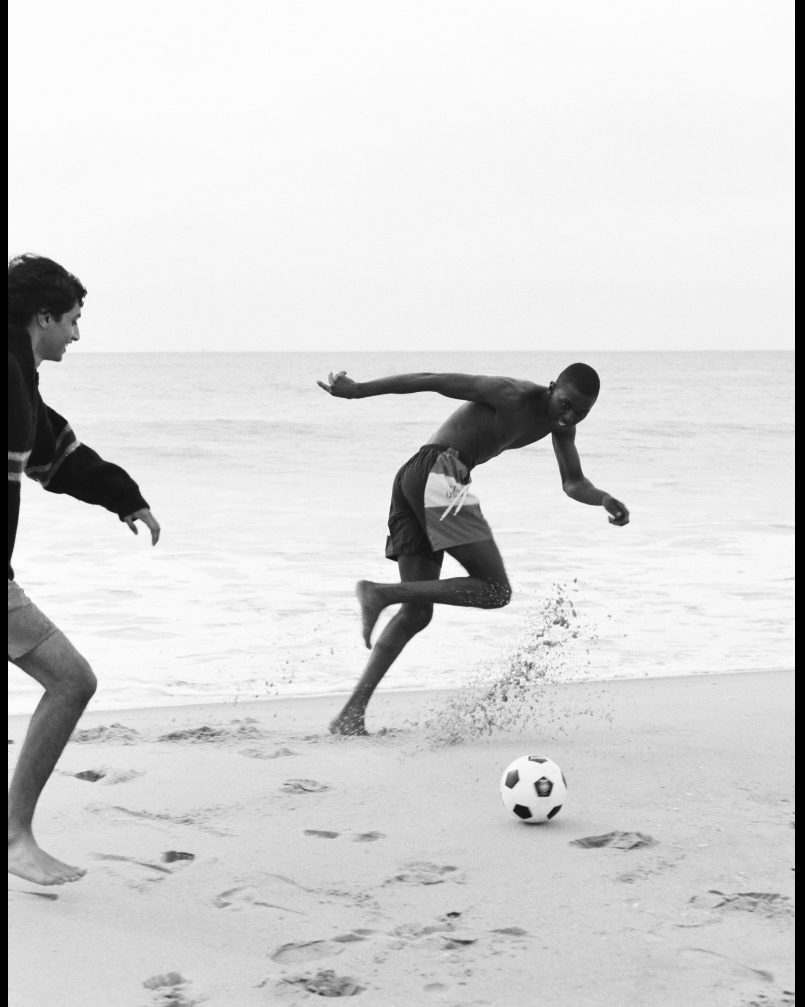 Two young men play soccer on a sandy beach, one barefoot and kicking the ball, with ocean and sky in the grayscale background.