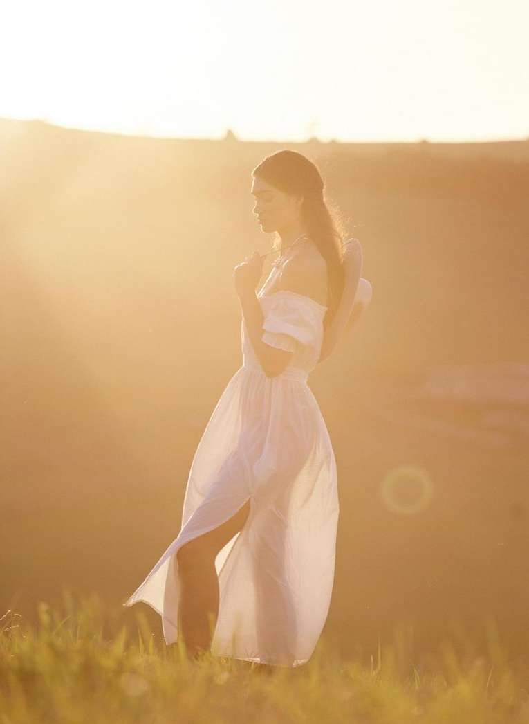 Woman in a long white dress standing in a grassy field during sunset by Ryan Brabazon