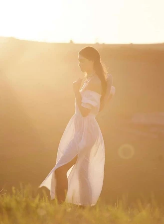 Woman in a long white dress standing in a grassy field during sunset by Ryan Brabazon