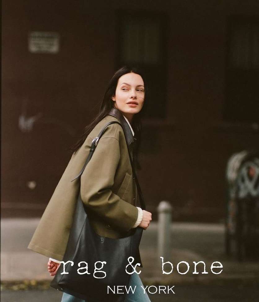 Woman in olive jacket with black tote bag walking on city street with brick building in the background, Rag & Bone branding.