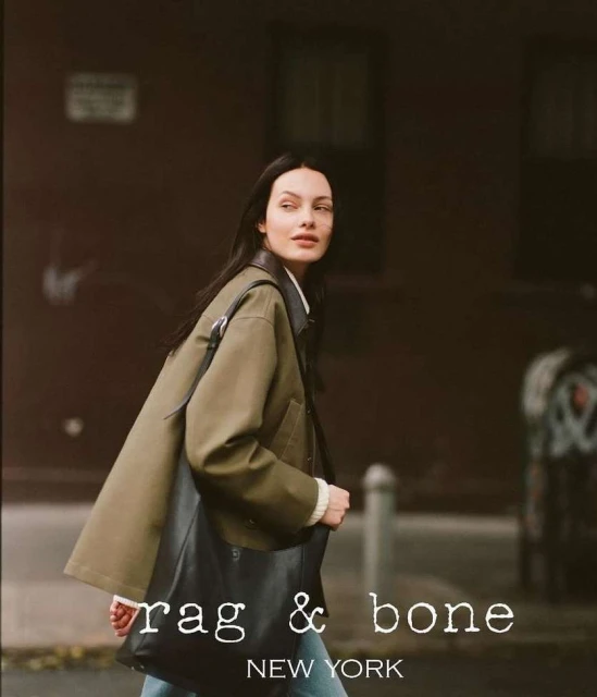 Woman in olive jacket with black tote bag walking on city street with brick building in the background, Rag & Bone branding.