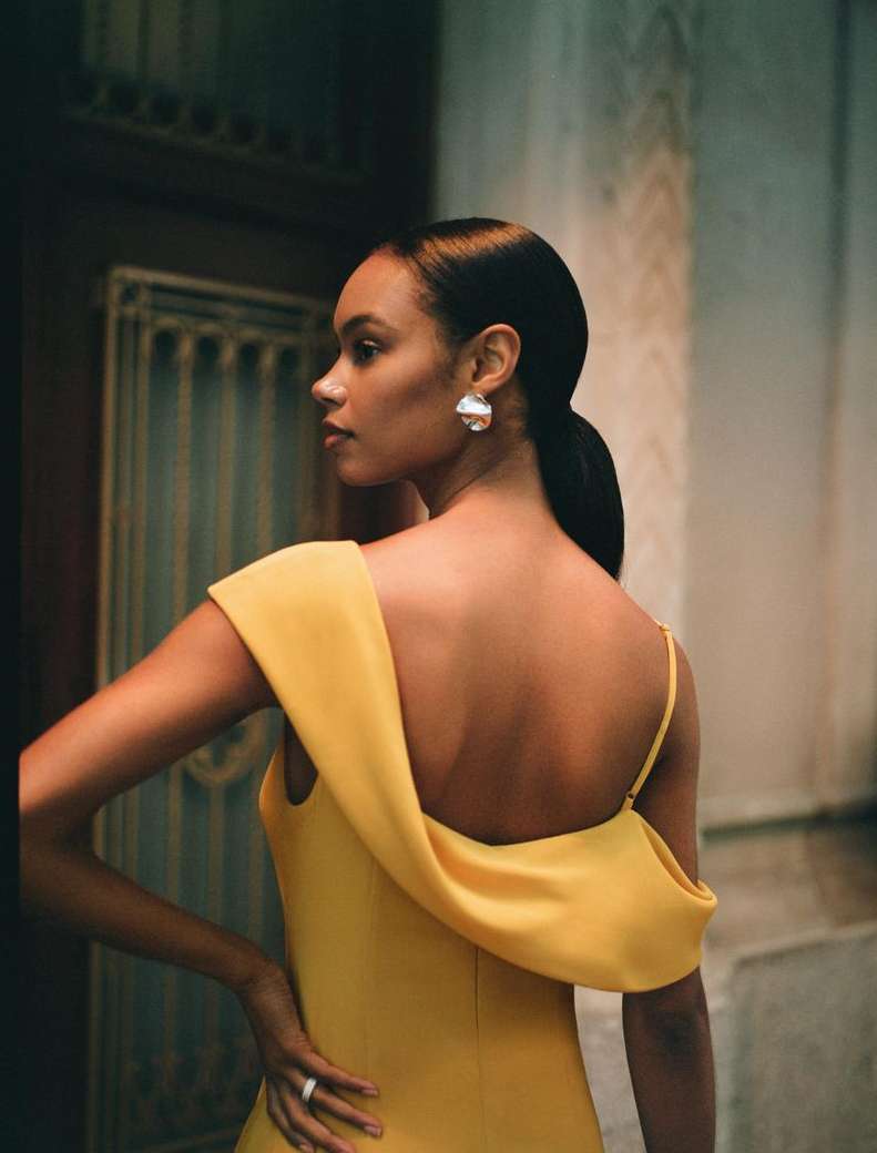Elegant woman in yellow dress poses in architectural setting, showcasing high fashion style and sophistication, enhanced by natural lighting.