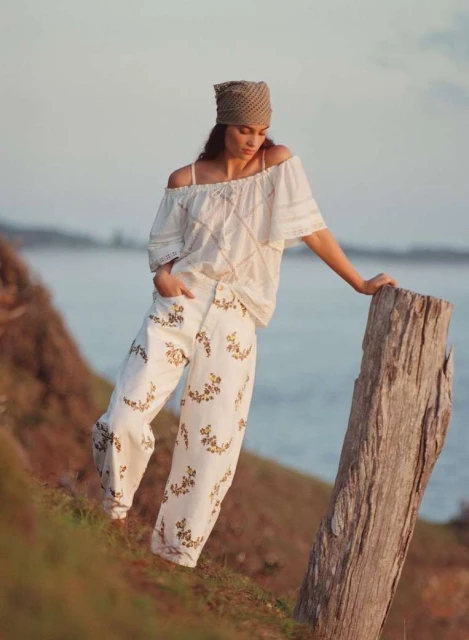 Adult woman poses in off-shoulder top and floral pants on a grassy hill near the sea, leaning against a weathered wooden post in natural light.