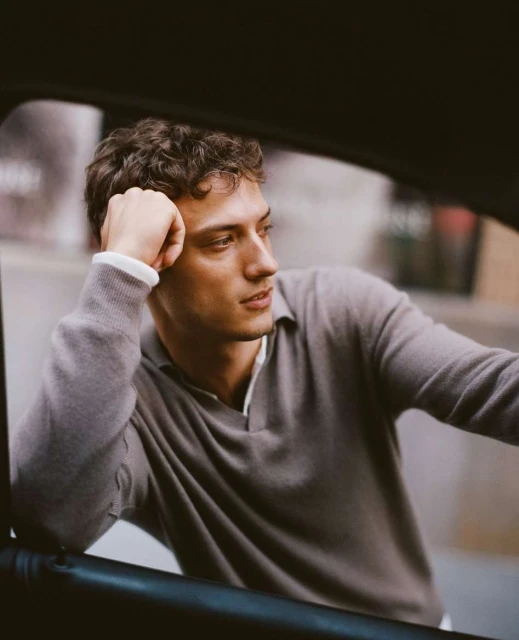 An adult male sits inside a car, resting his head on his hand. He wears a grey collared sweater and looks off to the side.