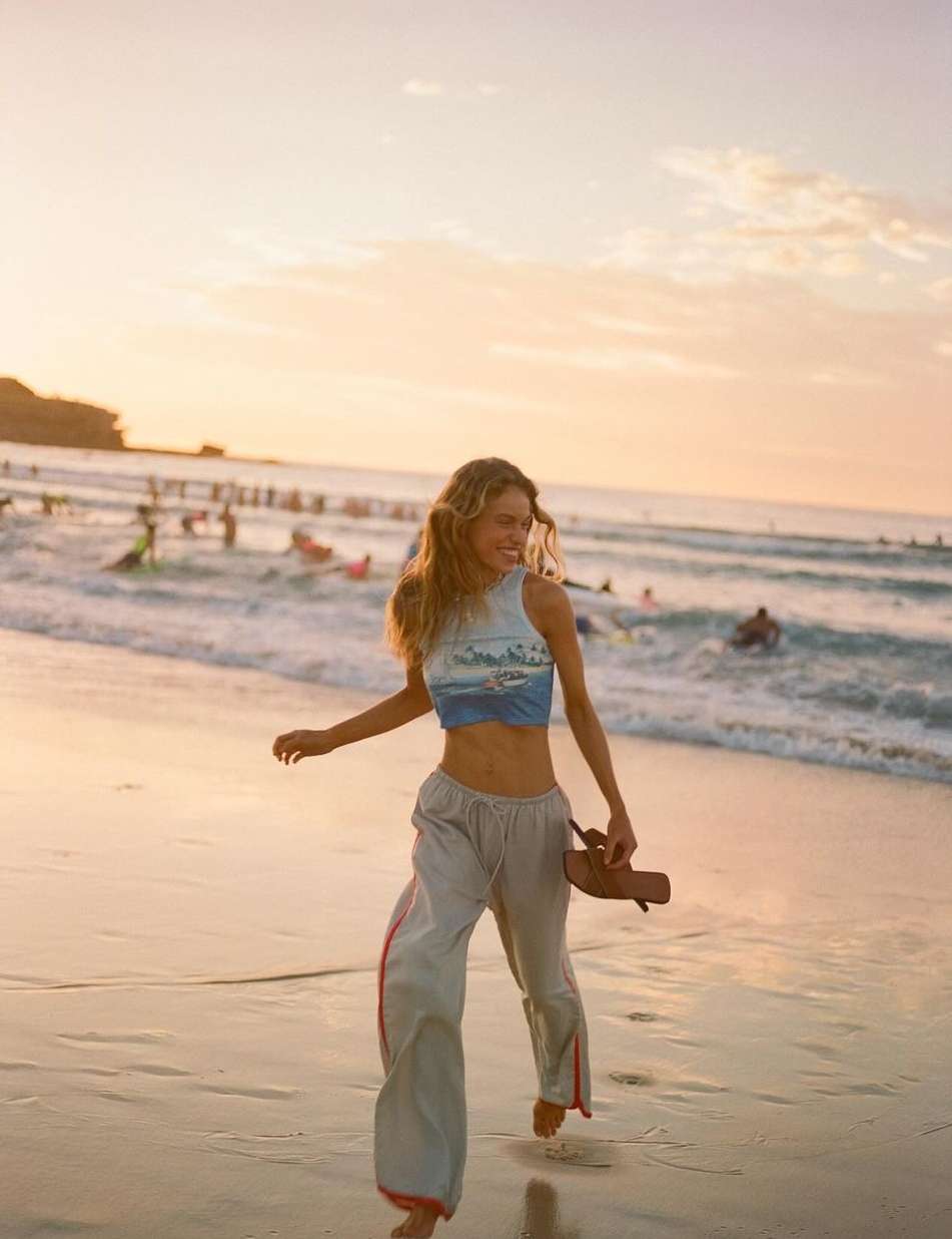 A smiling woman walks barefoot on a sandy beach at sunset, holding sandals. Waves and people in the water create a vibrant backdrop.
