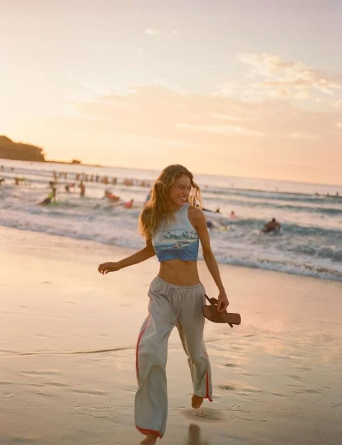 A smiling woman walks barefoot on a sandy beach at sunset, holding sandals. Waves and people in the water create a vibrant backdrop.