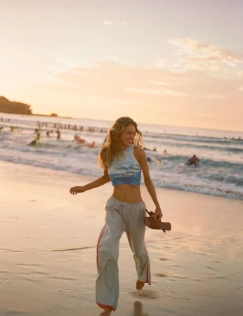 A smiling woman walks barefoot on a sandy beach at sunset, holding sandals. Waves and people in the water create a vibrant backdrop.