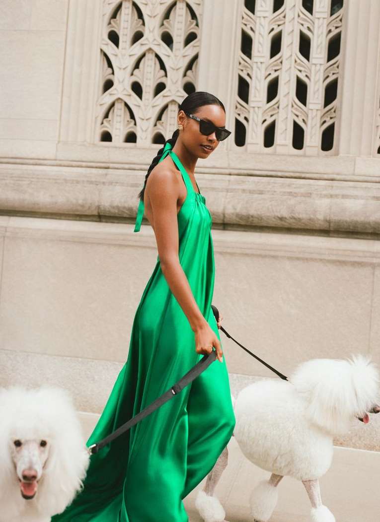 Elegant woman in flowing green gown walks two groomed white poodles against a light stone wall, showcasing fashion and canine companionship.