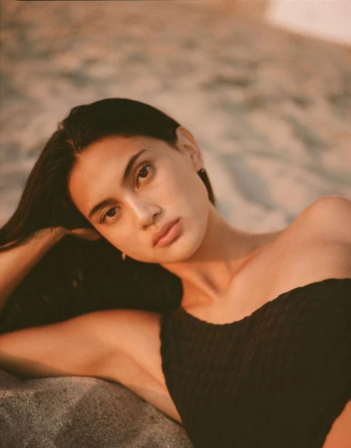 Young woman in a black swimsuit poses on the beach, illuminated by warm, golden light during the late afternoon or early evening.