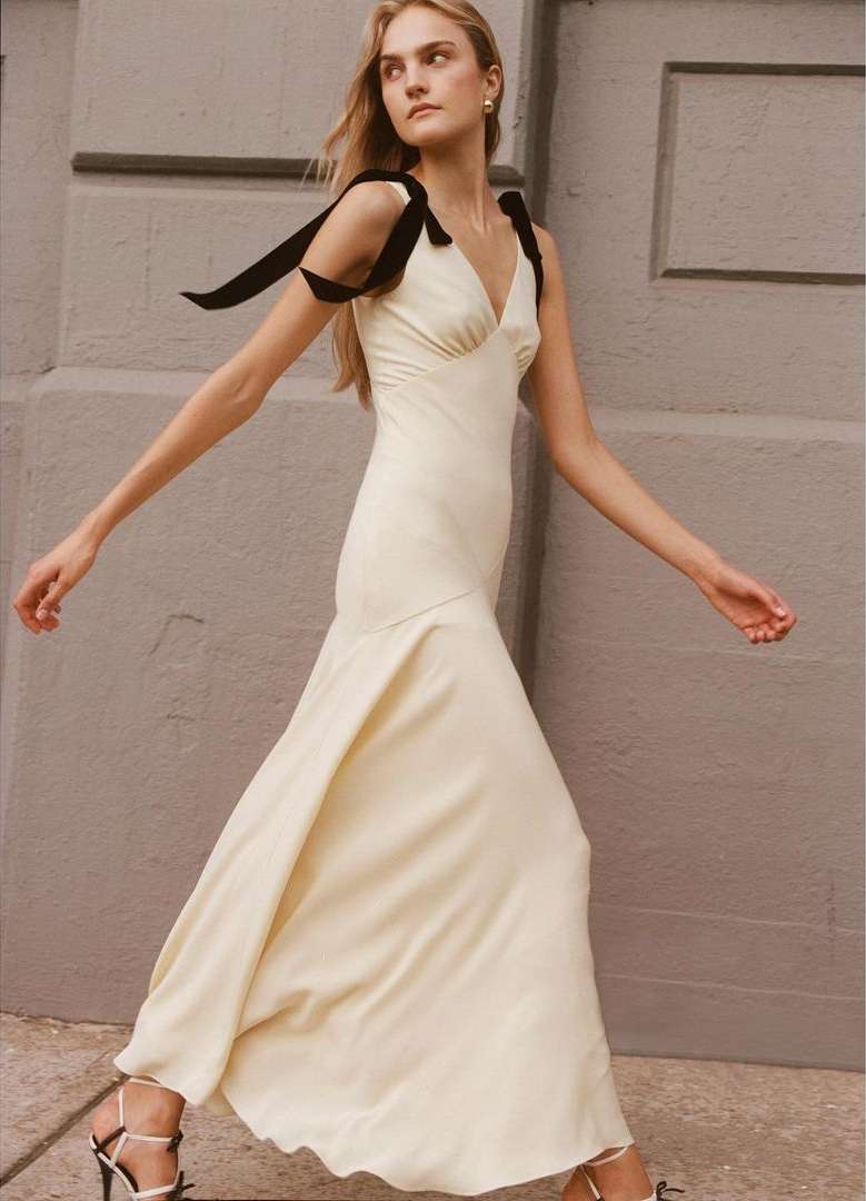 Elegant woman in a cream-colored dress with black ribbon straps walks past a gray wall in natural light, showcasing high-fashion style.