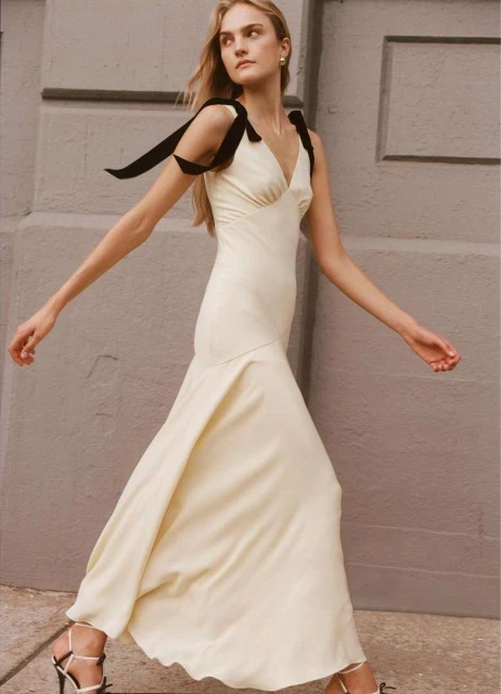 Elegant woman in a cream-colored dress with black ribbon straps walks past a gray wall in natural light, showcasing high-fashion style.