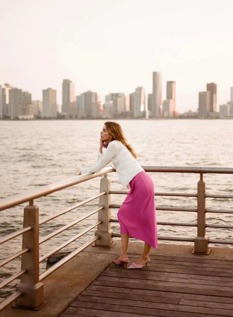 Woman in pink skirt and white sweater leans on pier railing with city skyline across the water in background, enjoying sunset.