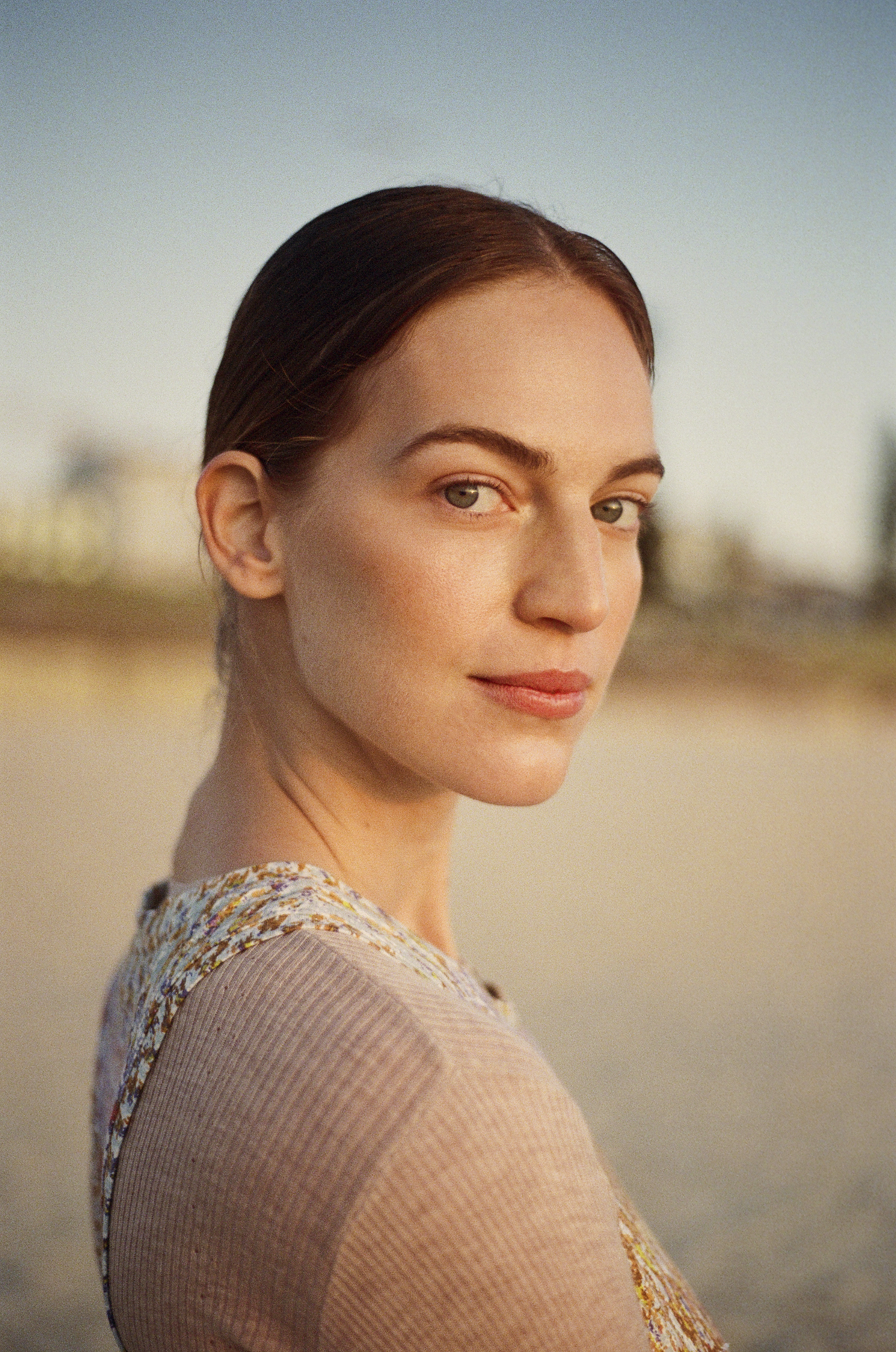 Close-up portrait of a woman with auburn hair wearing a floral and beige top, looking over her shoulder in outdoor setting with blue sky.