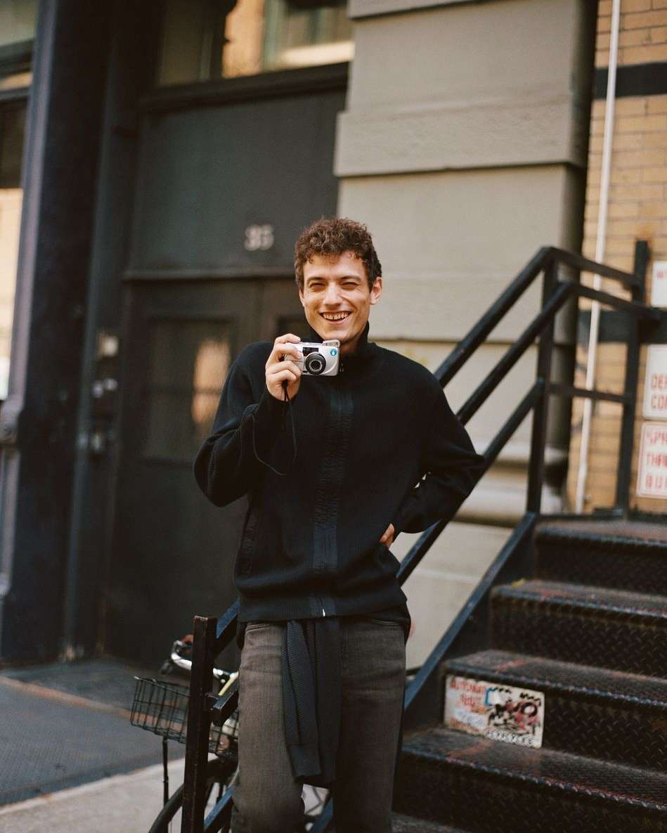 Young man with curly hair stands outside holding a digital camera, smiling on stairs next to a brick building with a metal staircase.